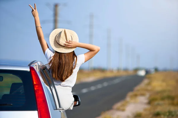 Mujer hermosa delgada en sombrero de paja disfrutando de viaje por carretera en un día de verano. Emocionada joven mujer levantando la mano con la señal de victoria de la ventana del coche. Chica montando sentado en la puerta del coche y asomándose por la ventana . —  Fotos de Stock