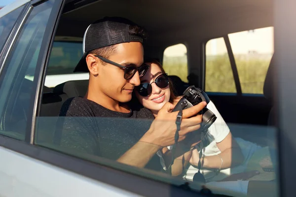 Blogger en gafas mostrando fotos a su novia en cámara profesional en viaje por carretera. Hombre fotógrafo revisar imágenes y examina el vídeo para el blog y vlog. Pareja multiracial viajan en coche . —  Fotos de Stock