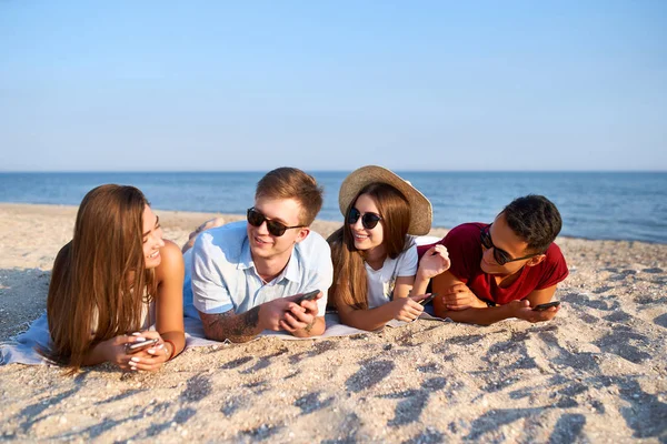 Молодая многонациональная группа тысячелетий отдыхает на пляжном полотенце у моря на белом песке. Стильные друзья висят на берегу, разговаривают, загорают со смартфонами в руках. Всегда подключенное поколение . — стоковое фото