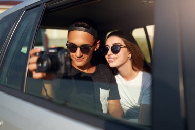 Gözlük Blogger yolda profesyonel kamera onun seyahat vlog filme. Adam fotoğrafçı kız arkadaşı ile açık pencereden fotoğraf çekmek yakın. Arka oto koltuğunda Multiracial hipsters çift.