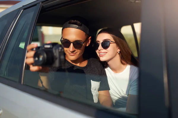 Blogger w okularach filmowanie jego podróży Vlog na profesjonalny aparat na wycieczkę. Człowiek fotograf robienia zdjęć z otwartego okna z dziewczyną w pobliżu. Para hipsters wielorasowe na tylnym siedzeniu samochodu. — Zdjęcie stockowe