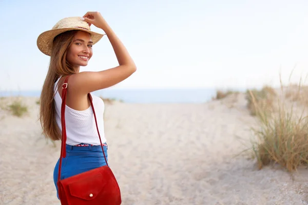 세련된 여성은 밀짚 모자, 여름 데님 반바지와 빨간색 유행 가방에 해변에 걸어. 매력적인 무두질 여성은 해안에 그녀를 따라 초대합니다. 웃는 소녀는 바다로 간다. 카피 스페이스. — 스톡 사진