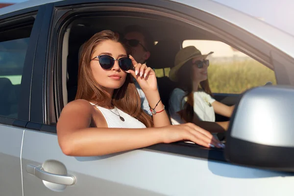 Группа друзей арендовала автомобиль в летнюю поездку и прибыла на морской пляж. Женщина в очках смотрит в окно машины. Пассажирка развлекается с друзьями в машине. Туристический стиль . — стоковое фото