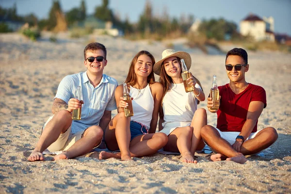 Группа друзей веселится, наслаждаясь напитком и отдыхая на пляже на закате в замедленной съемке. Молодые мужчины и женщины пьют пиво сидя на песке в теплый летний вечер . — стоковое фото