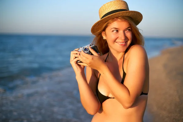 Young authentic woman with vintage retro film camera enjoying tropical beach on summer vacation. Female travel photographer in straw hat taking photos having fun at sea. Real girl unretouched shape. — Stock Photo, Image