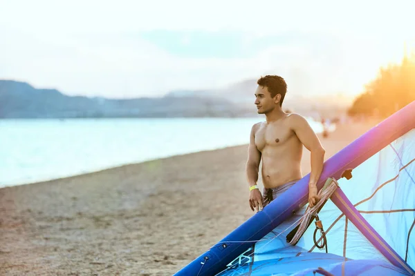 Uomo che tiene un aquilone in mano in piedi sulla spiaggia al tramonto dopo una buona sessione di kiteboarding. Kitesurfer con ala gode di kitesurf sull'isola tropicale. atletico ragazzo con abs andando a sgonfiare aquilone . — Foto Stock