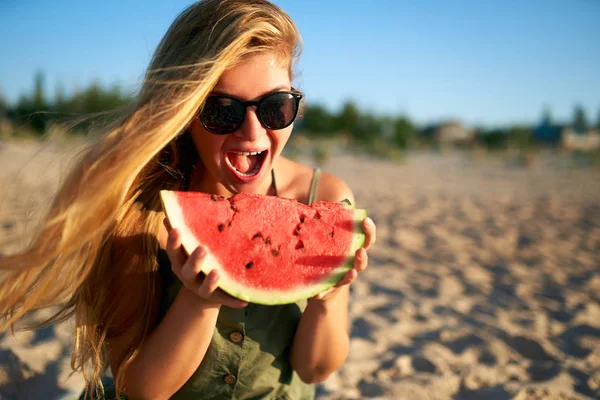 Happy young woman in glasses eating watermelon on the sandy beach on vacation. Girl joyfully holding fresh watermelon skip in her hands and have fun with hair fluttering in the wind. Youth lifestyle. — Stock Photo, Image