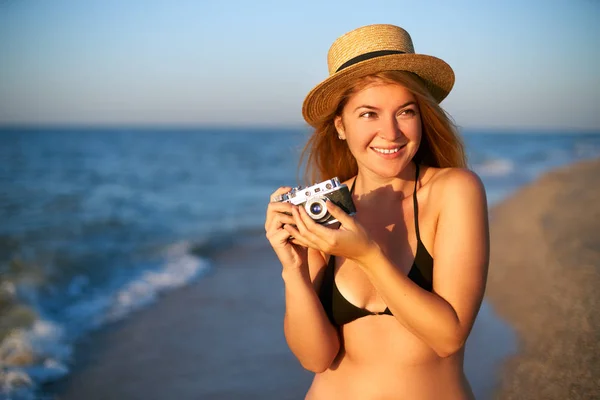 Young authentic woman with vintage retro film camera enjoying tropical beach on summer vacation. Female travel photographer in straw hat taking photos having fun at sea. Real girl unretouched shape. — Stock Photo, Image