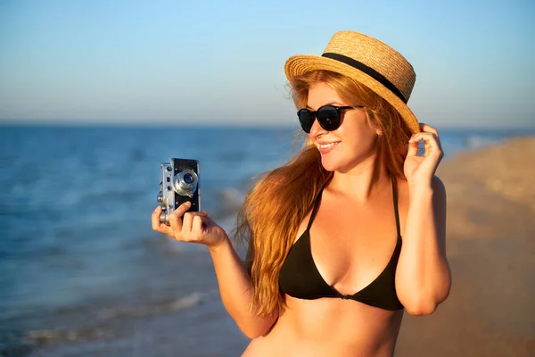 여름 방학에 열대 해변을 즐기는 빈티지 복고풍 필름 카메라와 젊은 정통 여성. 밀짚 모자와 비키니 수영복을 입은 여성 여행 사진작가가 해변에서 즐거운 사진을 찍고 있습니다.. — 스톡 사진