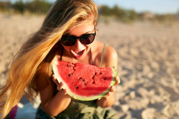 Happy young woman in glasses eating watermelon on the sandy beach on vacation. Girl joyfully holding fresh watermelon skip in her hands and have fun with hair fluttering in the wind. Youth lifestyle. — Stock Photo, Image