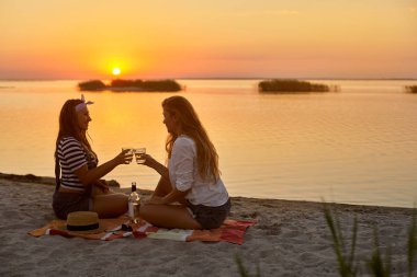 Kadınlar sahilde beyaz şarap ile gözlük Clink. Kız amigo tatil kutlamak ve tropikal adada tost ilan. Günbatımı deniz yakınında alkollü içecek içecek ile romantik piknik.