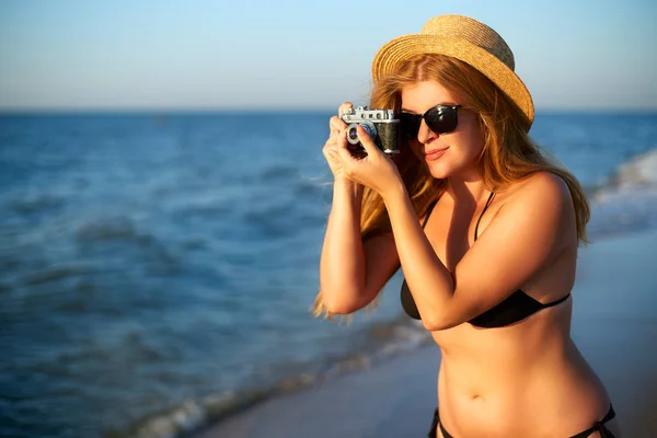 Young authentic woman with vintage retro film camera enjoying tropical beach on summer vacation. Female travel photographer in straw hat and bikini swimsuit taking photos having fun at the seaside. — Stock Photo, Image