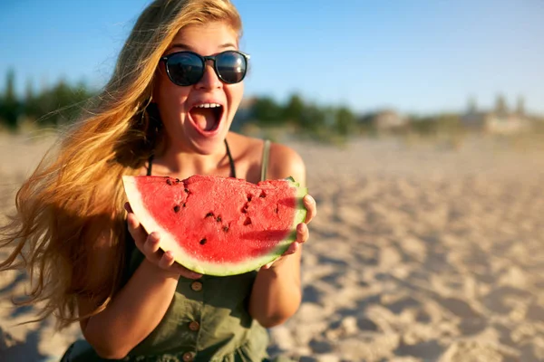 Happy young woman in glasses eating watermelon on the sandy beach on vacation. Girl joyfully holding fresh watermelon skip in her hands and have fun with hair fluttering in the wind. Youth lifestyle. — Stock Photo, Image