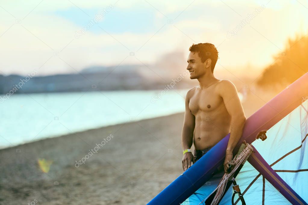Man holding a kite in hands standing at the beach on sunset after a good kiteboarding session. Kitesurfer with wing enjoys kitesurfing on tropical island. Athletic guy with abs going to deflate kite.