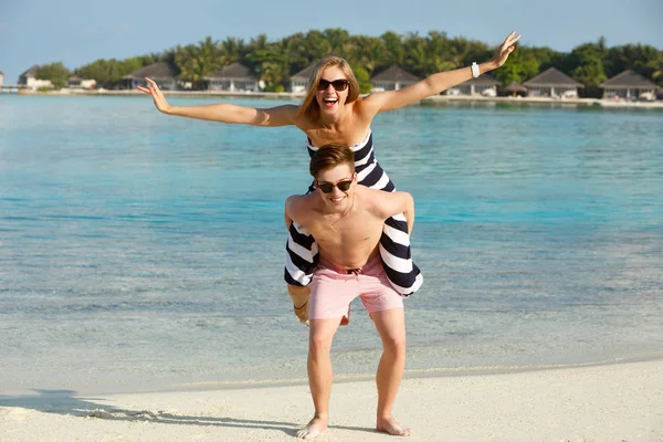 Glückliche junge schöne Paar haben Spaß und entspannen am Strand. Mann hält Frau mit Flugzeug auf dem Rücken. Bungalows mit Wellness-Resort, Palmen und blauer Lagune auf dem Rücken. Familienreisekonzept — Stockfoto