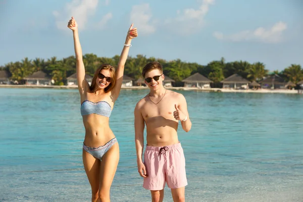 Feliz casal jovem se divertir e relaxar na praia. Homem e mulher mostram os polegares desfrutando de férias perto do oceano. Bungalows de estância termal, palmas e lagoa azul no fundo. Conceito de viagem familiar — Fotografia de Stock