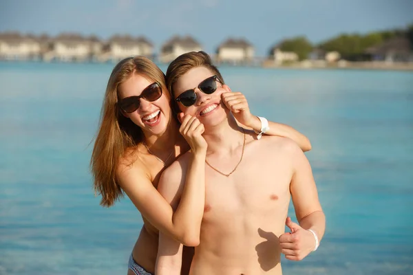 Feliz casal jovem se divertir e relaxar na praia. Mulher faz o homem sorrir segurando bochechas com as mãos. Bungalows de estância termal, palmas e mar azul no fundo. Conceito de viagem familiar — Fotografia de Stock