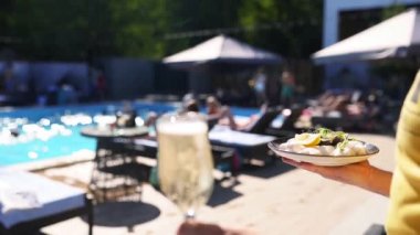 Garson tepside istiridye ve bir bardak şampanya ya da lüks bir otelde ya da plaj kulübünde havuz kenarında köpüklü şarap taşıyor. Tatil köyünde yavaş çekimde deniz ürünleri ve içecekler servis eden adam..