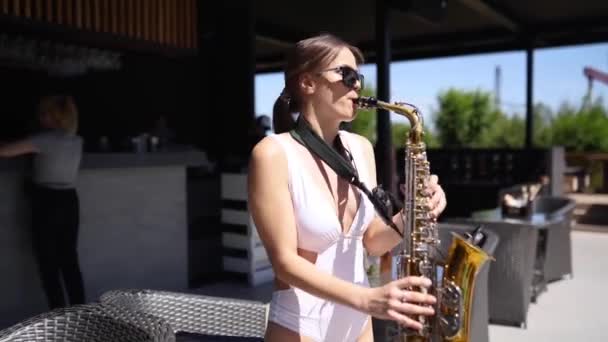 Attractive saxophonist lady is playing on saxophone near pool at beach club. Pretty sax girl musician in hot white bikini dances and plays on weekend party onhot summer day. — Stock Video