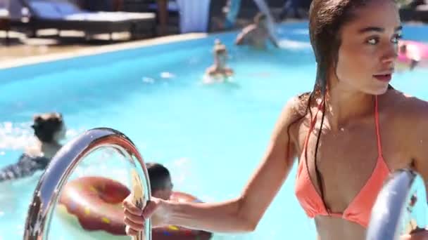 Mladá atraktivní mokrá běloška v oranžových bikinách vychází z bazénu v beach clubu. Hezká dívka opouští bazén podle kovového žebříku ve zpomaleném filmu v horkém letním dni. — Stock video