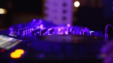 DJ, disko kulübünde müziği karıştırmak için ses konsolunu kontrol ediyor. Parmaklar düğmelere basar, güvertede elektronik müzik çalar. Gece kulübü dans partisinde renkli ışıklar gösteriliyor. Ağır çekimde kapat..