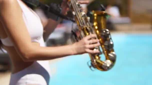 Attractive saxophonist lady is playing on saxophone near pool at beach club. Pretty sax girl musician in hot white bikini dances and plays on weekend party onhot summer day. — Stock Video