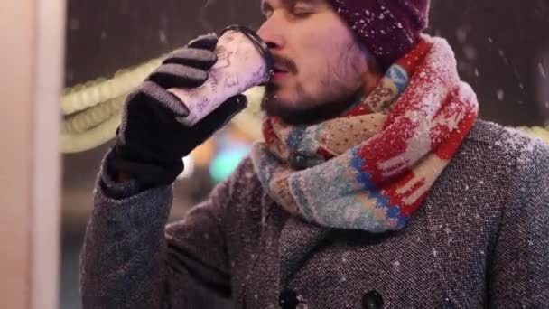 Baard stijlvolle man in grijze jas en gebreide sjaal drinkt koffie uit papieren beker en kijkt naar de camera glimlachend. Kerstmarkt en Nieuwjaar winterbeurs op achtergrond. Sneeuwnacht. Wintermode. — Stockvideo