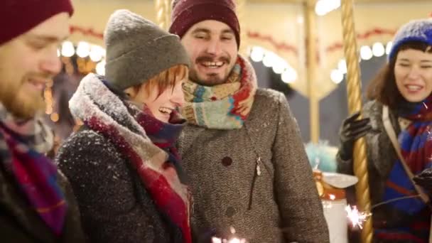 Amigos sonrientes se divierten con bengalas montando carrusel en el mercado de Navidad. Hombres y mujeres felices colgando en el parque de atracciones de la feria de invierno de Año Nuevo. Luces de guirnalda en el fondo. La gente se relaja en la noche nevada — Vídeos de Stock