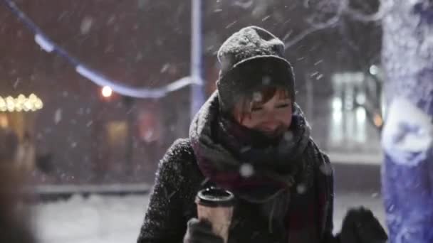 Grupo de amigos felices caminando bajo las nevadas con tazas de café de papel en las manos y divertirse jugando con la nieve. La gente se calienta con bebidas calientes y cuelga en la noche de invierno. Navidad y Año Nuevo . — Vídeos de Stock