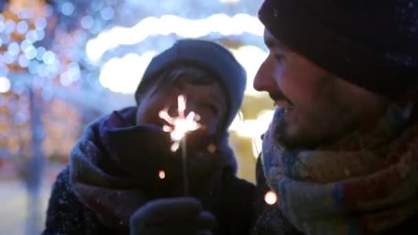 Gelukkige vrienden wandelen met sterretjes in handen en hebben plezier op besneeuwde winternacht in feestelijke verlichtingstunnel. Mensen hangen op de kerstmarkt. slingers, carrousellampjes op achtergrond. — Stockvideo