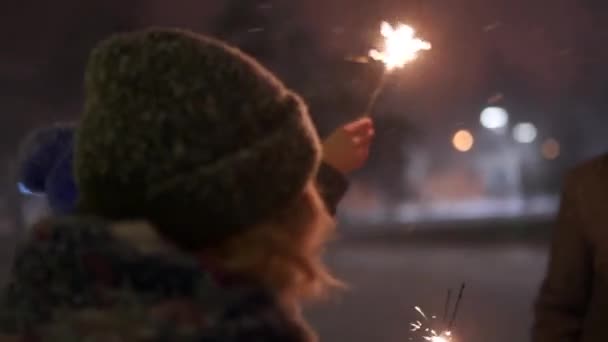 Gli amici allegri si divertono con scintille in mano. La gente balla la notte d'inverno con nevicate, mercatino di Natale, ghirlande di lampade sullo sfondo al rallentatore all'aperto. Capodanno, Festa di compleanno . — Video Stock