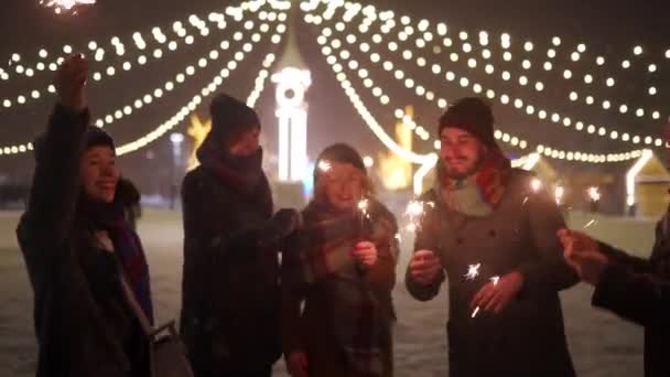 Teman-teman ceria bersenang-senang melambaikan tangan dengan kembang api di tangan. Malam Natal, Liburan Tahun Baru atau Pesta Ulang Tahun. Happy people jump partiying on winter night with snow and lamp garlands on background . — Stok Video