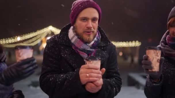 Amigos felizes saindo e beber bebidas quentes. As pessoas torcem copos de papel desfrutando de café no café ao ar livre na neve fria da noite de inverno em câmera lenta. Natal, conceito de feriados de Ano Novo . — Vídeo de Stock