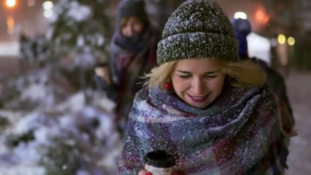 Groep van gelukkige vrienden wandelen onder sneeuw met papieren koffiekopjes in handen en veel plezier met het spelen met sneeuw. Mensen warmen op drinken warme dranken en hangen op kermis en nieuwjaarsmarkt — Stockvideo