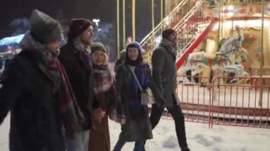 Friends walking under snowfall near carousel on snowy winter night., have fun and communicate. Group of happy people hanging at Christmas market. Christmas fair entertainment and New Year Holidays.