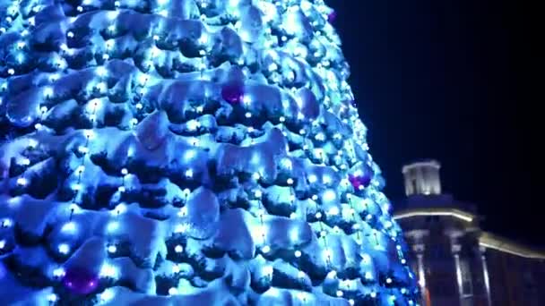 Nagy karácsonyfa díszített fényes koszorúk villogó a város főterén havas téli éjszakán. Fényképezőgép repül körül izzó újévi fa tele dekorációk a szabadban az utcán. Fényszóró. — Stock videók