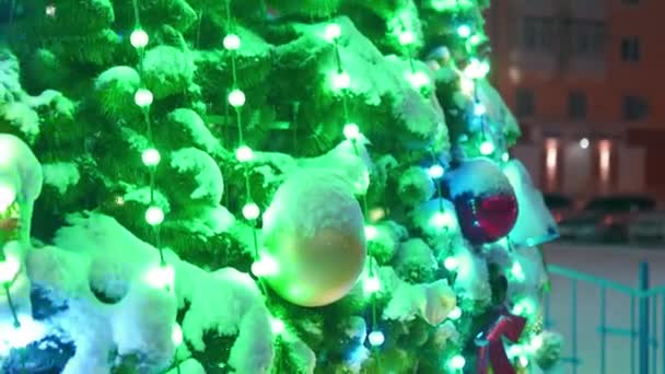 Grande árvore de Natal com guirlandas luminosas e decorações na praça da cidade na noite de inverno nevado. Close-up. A câmera voa ao redor da árvore de Ano Novo decorada brilhante cheia de decorações ao ar livre. Mostra de luz — Vídeo de Stock