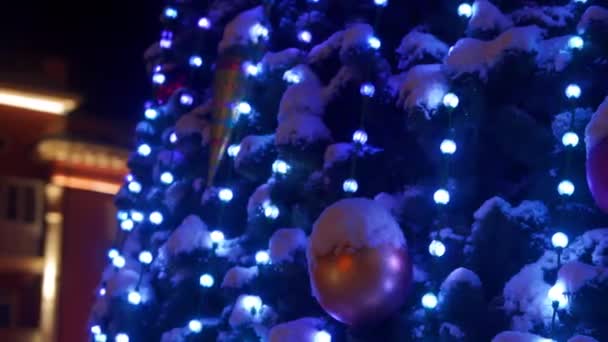 Gran árbol de Navidad con guirnaldas luminosas y decoraciones en la plaza de la ciudad en la noche nevada de invierno. Primer plano. Árbol de Año Nuevo decorado brillante lleno de decoraciones al aire libre. Izquierda derecha panorama . — Vídeos de Stock