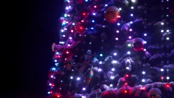 Μεγάλο χριστουγεννιάτικο δέντρο με φωτεινές γιρλάντες και διακοσμήσεις στην πλατεία της πόλης το χιονισμένο βράδυ του χειμώνα. Κοντινό πλάνο. Λαμπερό διακοσμημένο δέντρο Πρωτοχρονιάς σε εξωτερικούς χώρους. Δεξί πανόραμα. — Αρχείο Βίντεο