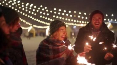 Bir grup arkadaş ellerinde maytaplarla el sallayarak eğleniyorlar. Mutlu insanlar kış gecesi kar yağışı, Noel pazarı ve arka planda lamba çelenkleriyle parti yapıyorlar. Yeni yıl tatili ya da doğum günü partisi.