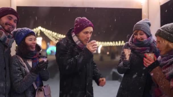 Happy friends hanging out and drink hot warming beverages. People cheer paper cups enjoying coffee in outdoor cafe on cold winter night snowfall in slow motion. Christmas, New Year holidays concept. — Stock Video