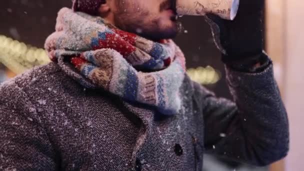 Bärtiger, stilvoller Mann in grauem Mantel und Strickschal trinkt Kaffee aus Pappbecher und blickt lächelnd in die Kamera. Christkindlmarkt und Neujahrswintermarkt im Hintergrund. verschneite Nacht. Wintermode. — Stockvideo