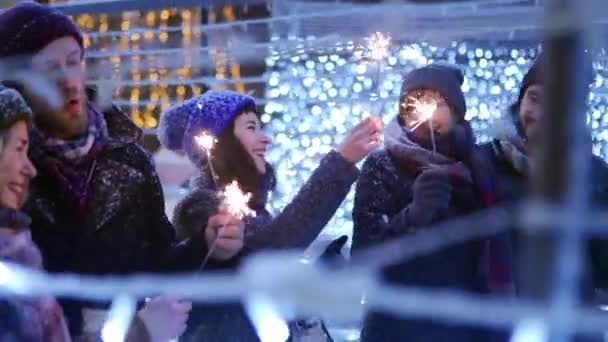 Gelukkige vrienden wandelen met sterretjes in handen en hebben plezier op besneeuwde winternacht in feestelijke verlichtingstunnel. Mensen hangen op de kerstmarkt. slingers, carrousellampjes op achtergrond. — Stockvideo