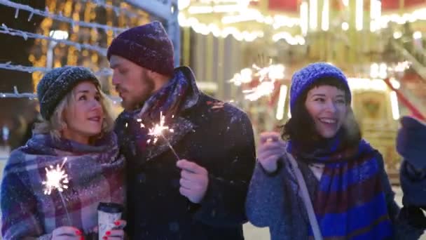 Gelukkige vrienden wandelen met sterretjes in handen en hebben plezier op besneeuwde winternacht in feestelijke verlichtingstunnel. Mensen hangen op de kerstmarkt. slingers, carrousellampjes op achtergrond. — Stockvideo