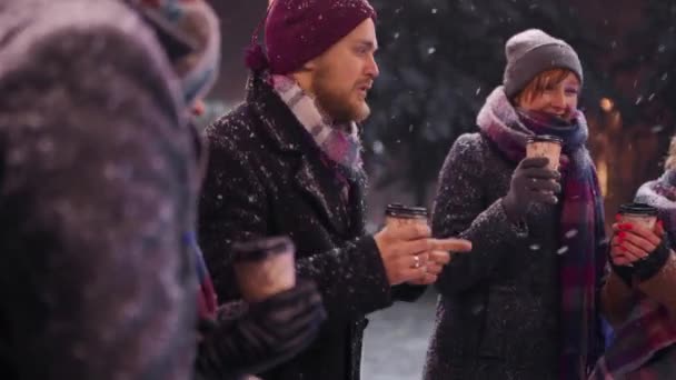 Amigos felices pasando el rato y beber bebidas calientes. La gente anima las copas de papel de vinilo caliente bailando en la cafetería al aire libre en la fría noche nevada de invierno en cámara lenta. Navidad, concepto de vacaciones de Año Nuevo . — Vídeos de Stock