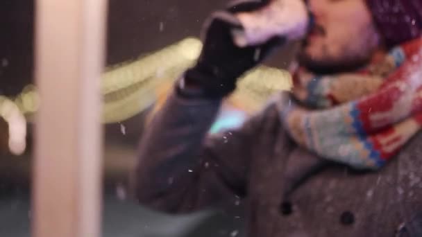 Γενειοφόρος κομψός άντρας με γκρι παλτό και πλεκτό φουλάρι πίνει καφέ από χάρτινο κύπελλο και κοιτάζει την κάμερα χαμογελώντας. Χριστουγεννιάτικη αγορά και Πρωτοχρονιά χειμερινή έκθεση στο παρασκήνιο. Χιονάτη νύχτα. Χειμερινή μόδα. — Αρχείο Βίντεο