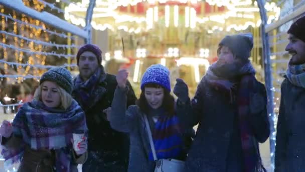 Amigos felices caminando con bengalas en las manos y divirtiéndose en la noche de invierno nevada en el túnel de iluminación festivo. Gente colgando en el mercado de Navidad. Lámparas de guirnalda, luces de carrusel sobre fondo . — Vídeo de stock