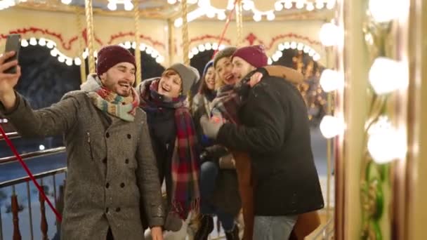 Happy people taking selfie photo on smartphone camera at New Year winter fair amusement park. Smiling friends have fun riding carousel at Christmas market. Blogger works. Garland lights on backdrop. — Stockvideo