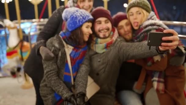 Szczęśliwi ludzie robiący selfie na aparacie fotograficznym smartfona w noworocznym parku rozrywki Winter Fair. Uśmiechnięci przyjaciele dobrze się bawią jeżdżąc na karuzeli na jarmarku. Bloger działa. Światła girlandowe na tle. — Wideo stockowe
