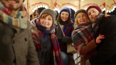 Mutlu insanlar Yılbaşı Fuarı eğlence parkında akıllı telefon kamerasıyla selfie çekiyorlar. Gülümseyen arkadaşlar Noel pazarında atlı karınca sürerken eğlenirler. Blogcu çalışıyor. Arka planda çelenk ışıkları.